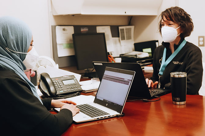 two women in masks sitting across from each other at a desk with laptops